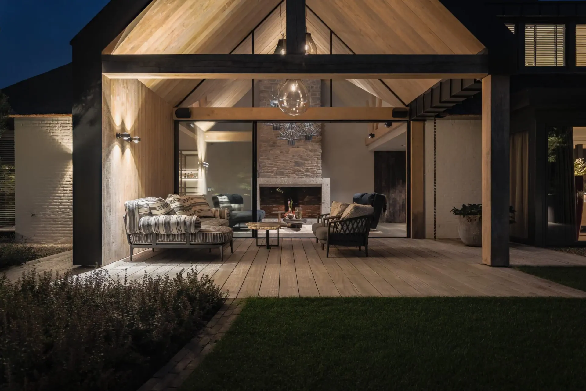 Nachtzicht van een luxe moderne overkapping ontworpen door Eric Kant, met sfeervolle verlichting op het houten plafond, een houten terras en zicht op de verlichte natuurstenen haard binnen.