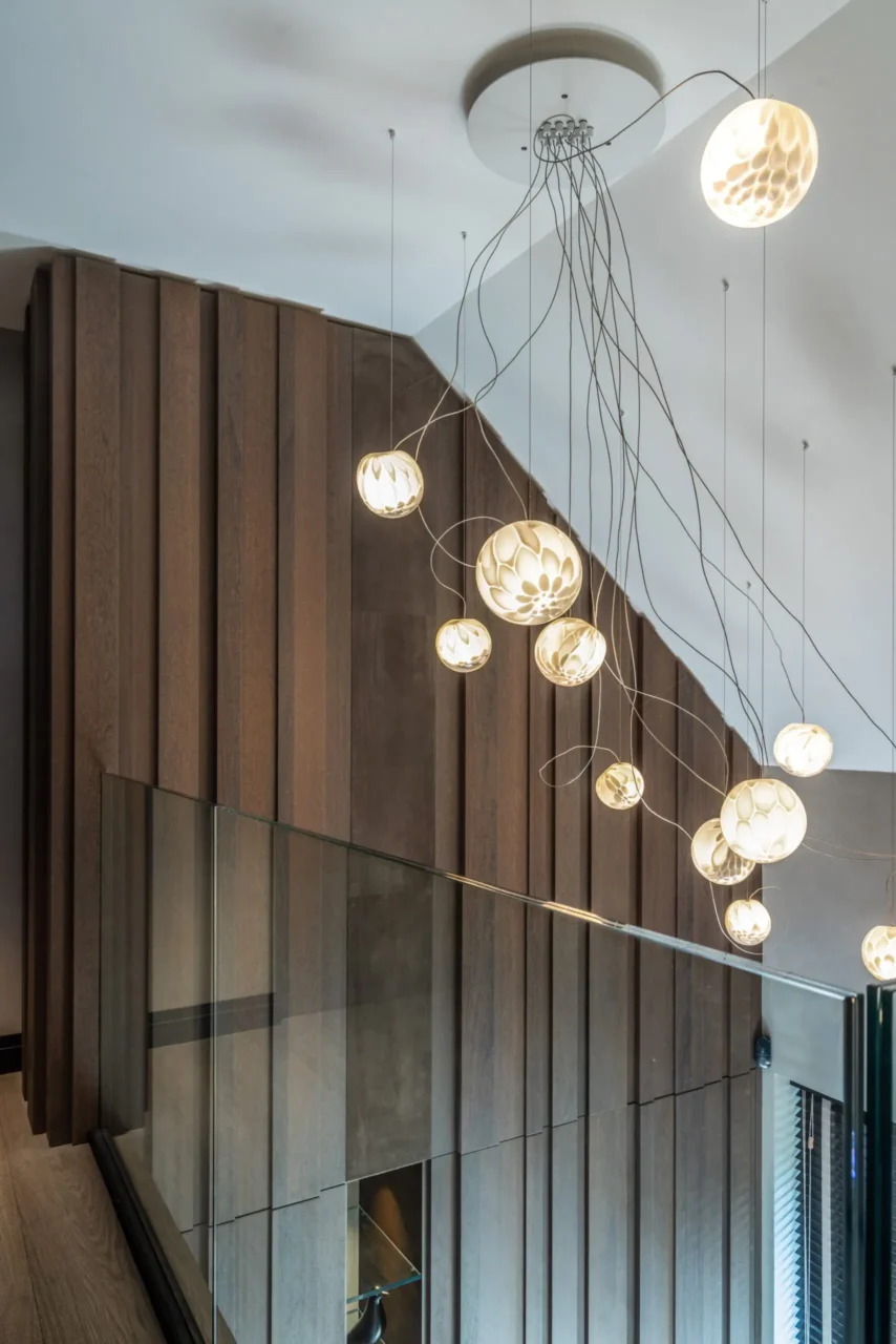 Detail van de hal en vide in Villa Essenza door ERIC KANT met een imposante wand van houten latten, een artistieke glazen hanglamp in cascadevorm en een grote passpiegel die de ruimte reflecteert.