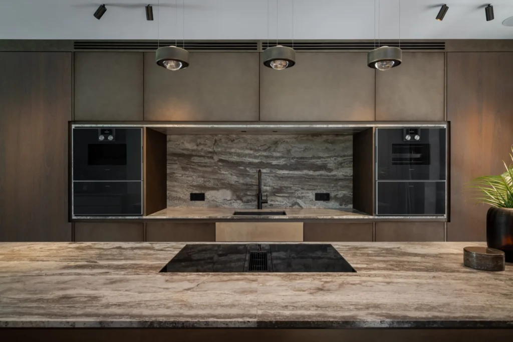 Frontaal zicht op de high-end keuken in Villa Essenza door ERIC KANT met een travertijn achterwand en eiland, geïntegreerde ovens en een strakke inductiekookplaat.