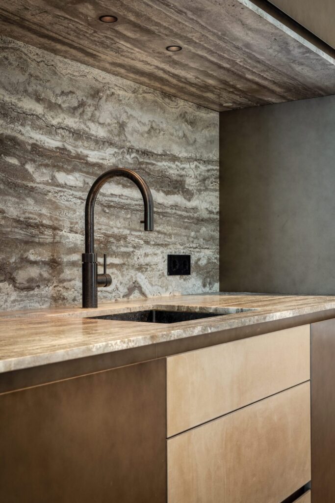 Detailopname van een travertijn aanrechtblad en achterwand in Villa Essenza door ERIC KANT met een zwarte designkraan en maatwerk kasten bekleed met een zachte textuur.