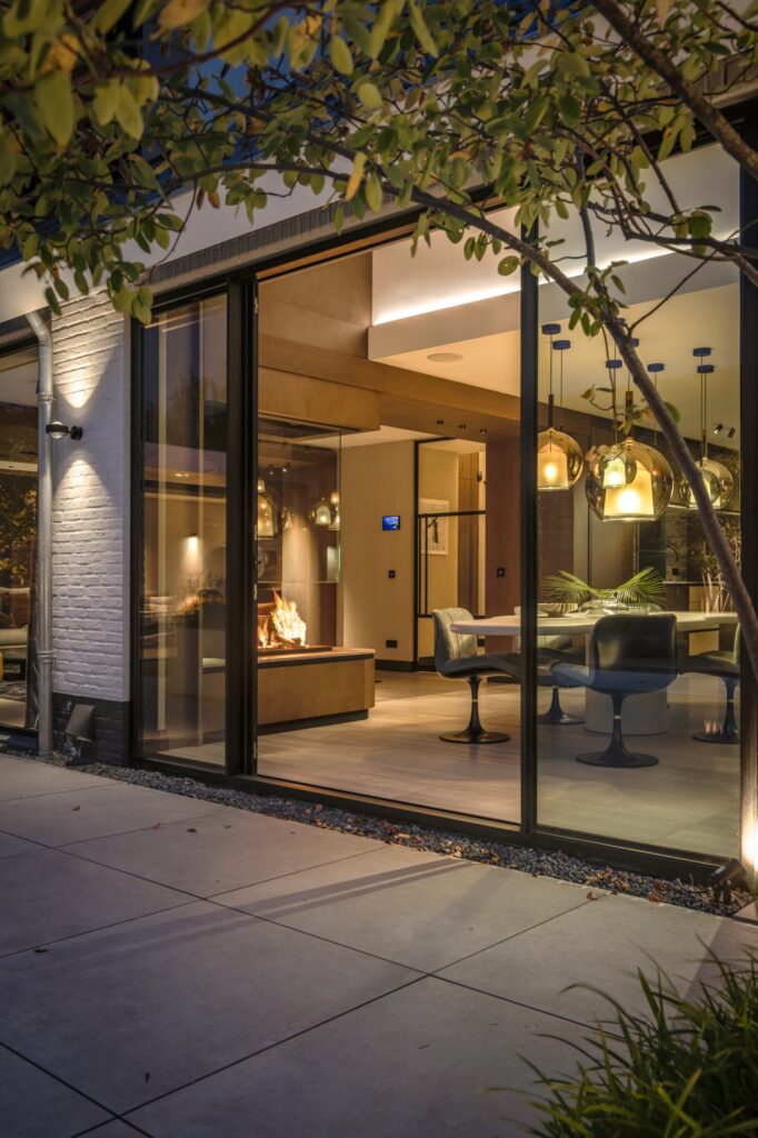 Avondzicht op de gevel van Villa Essenza door ERIC KANT met grote glazen schuifdeuren die zicht bieden op de verlichte eethoek, de designlampen en de brandende doorkijkhaard.