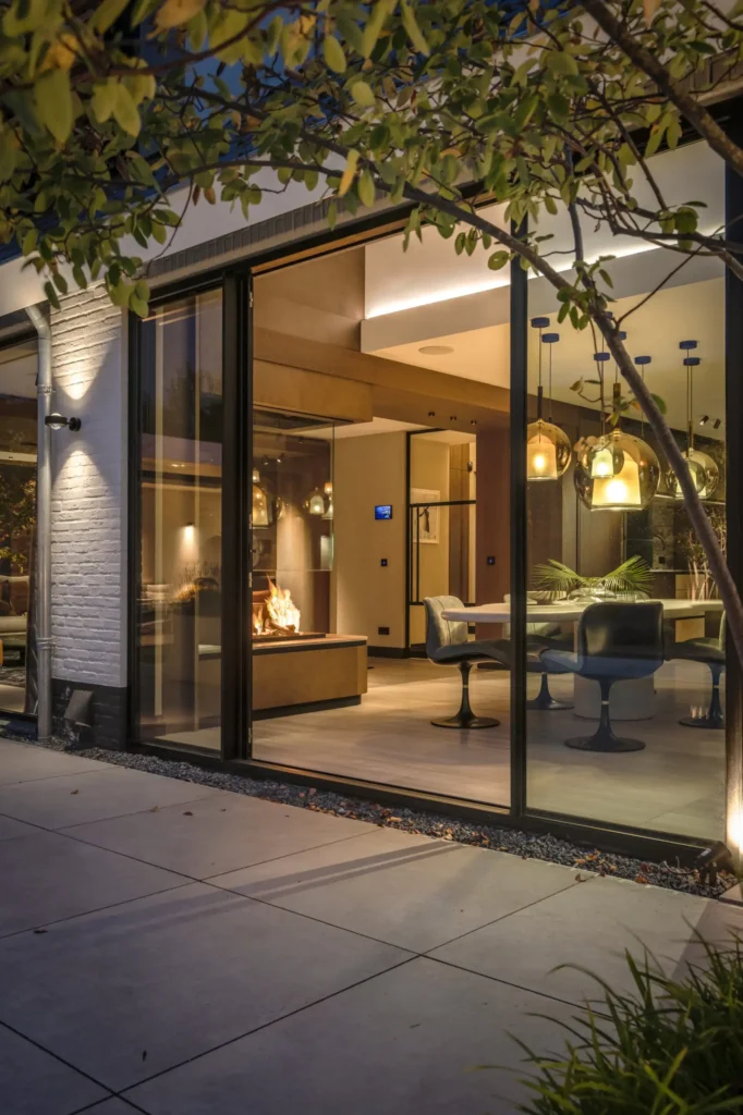 Avondzicht op de gevel van Villa Essenza door ERIC KANT met grote glazen schuifdeuren die zicht bieden op de verlichte eethoek, de designlampen en de brandende doorkijkhaard.