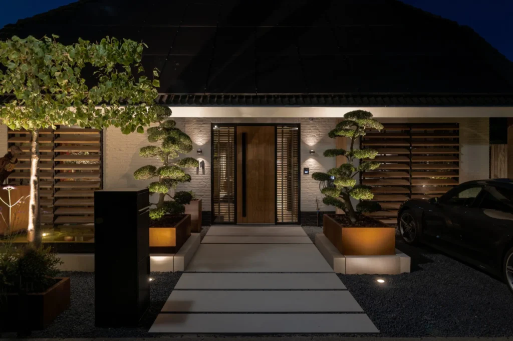 Vooraanzicht van de entree van Villa Essenza door ERIC KANT bij avond, met verlichte niwaki-bomen in cortenstalen bakken, een eikenhouten deur en een modern lijnenspel van licht en schaduw.
