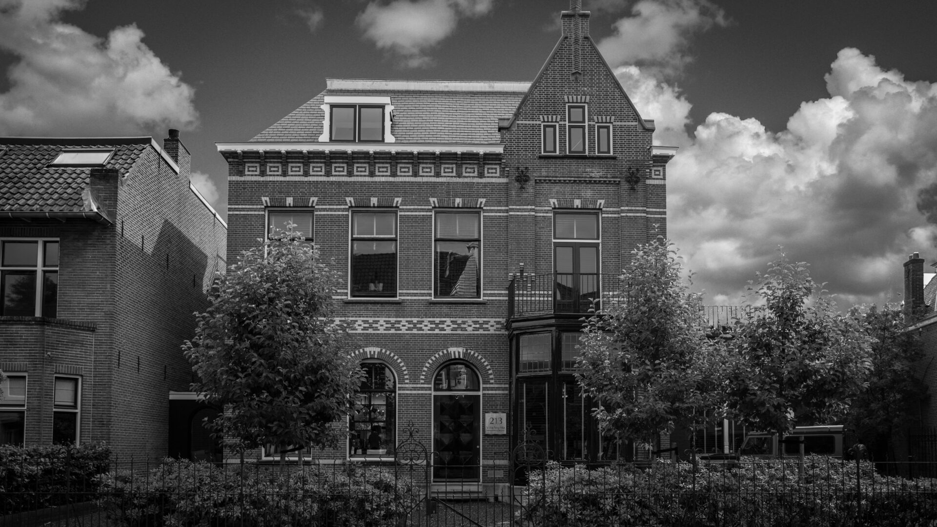 Een karakteristieke zwart-wit opname van het monumentale kantoorpand van ERIC KANT in Breda, met het naambordje zichtbaar naast de entree en een fraai aangelegde voortuin onder een bewolkte lucht.
