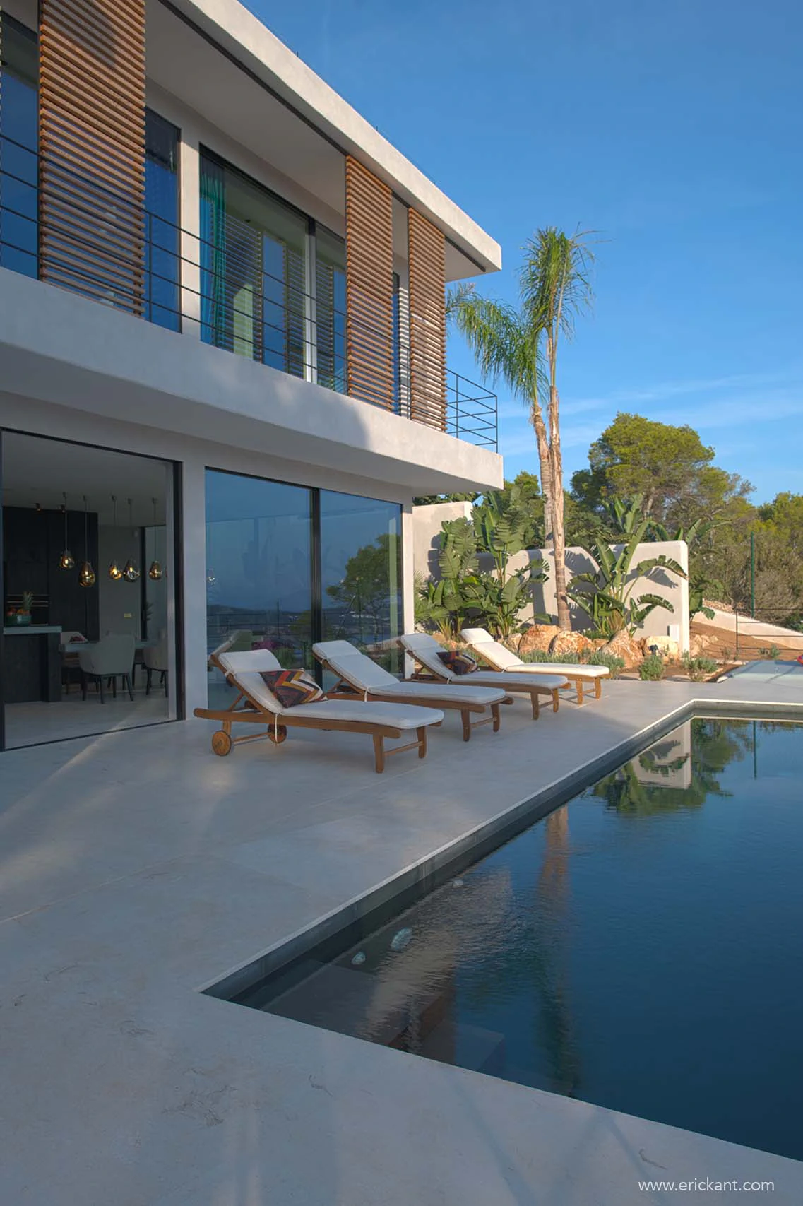 Een moderne luxe villa op Ibiza door ERIC KANT met een strak overloopzwembad, minimalistische houten shutters, kamerhoge glazen puien en een zonneterras met houten ligbedden