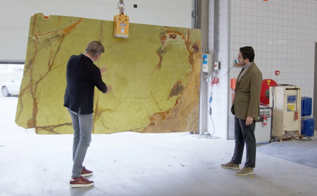 Ontwerper Eric Kant inspecteert een indrukwekkende plaat groen onyx natuursteen in een werkplaats, terwijl hij de unieke adering en gelaagdheid uitlegt aan een jongere collega.