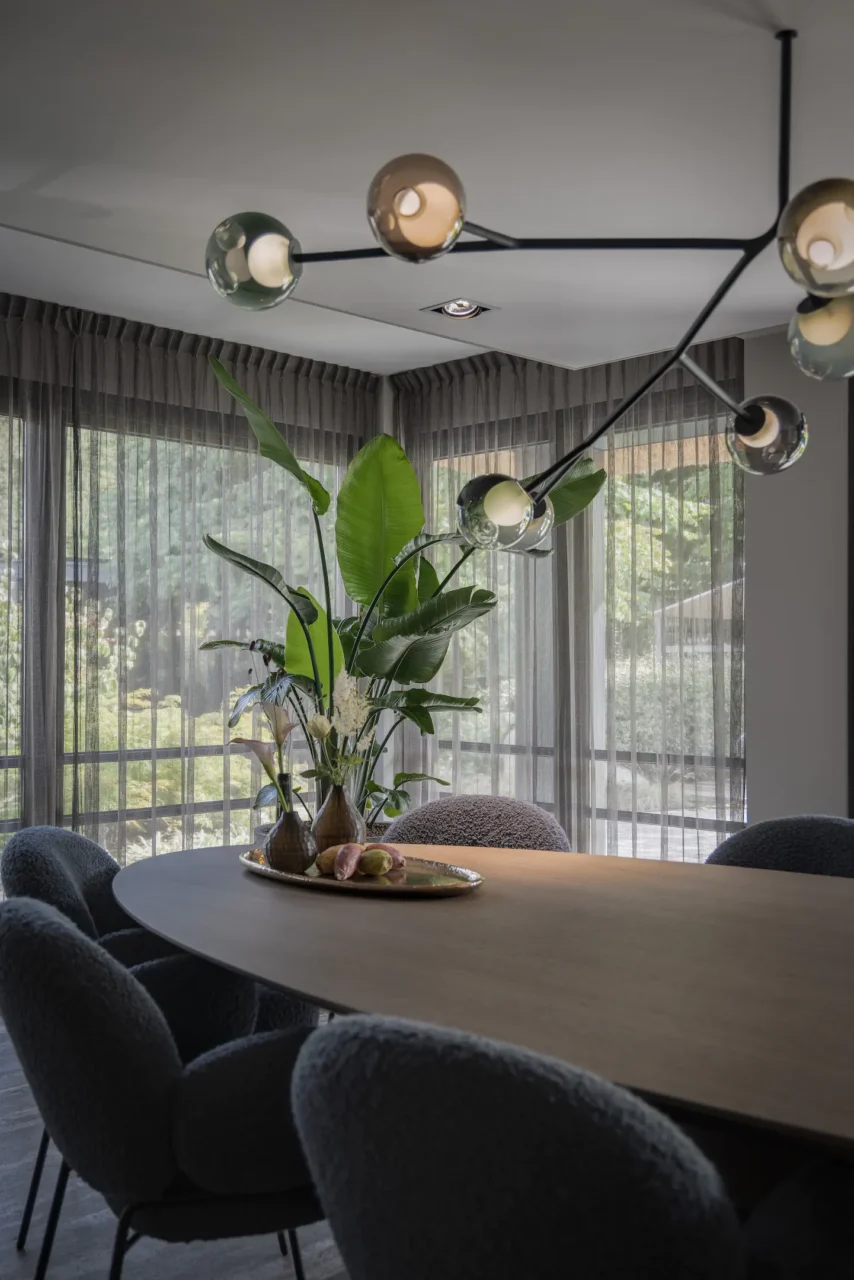 Een detailopname van een eethoek door ERIC KANT met een ovale houten tafel, zachte teddy-stof stoelen in antraciet, een moderne zwarte designlamp met glazen bollen en een grote Strelitzia plant voor kamerhoge vitrage.