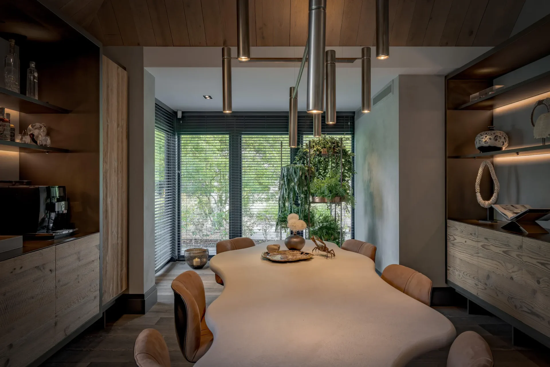Een luxe eetkamer ontworpen door ERIC KANT met een opvallende organisch gevormde eettafel, moderne cilinderverlichting, maatwerk kastenwanden van hout en brons, en zicht op de tuin door grote ramen met lamellen.