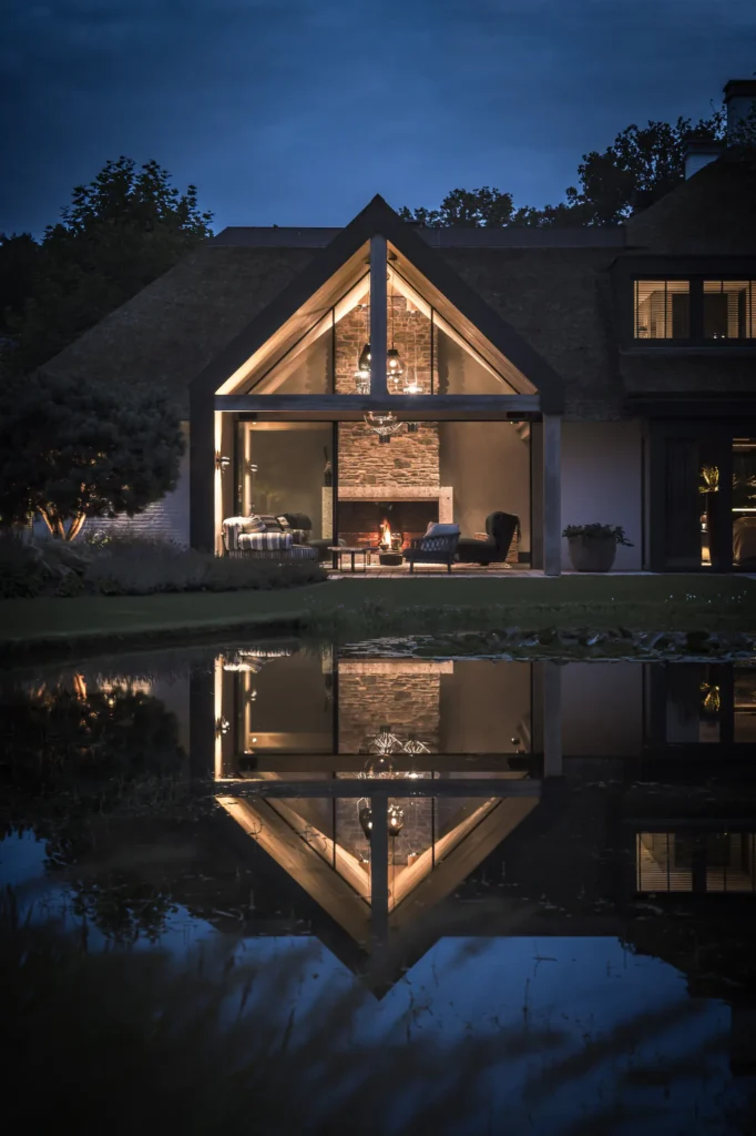 Een verlichte, rietgedekte villa bij avond door ERIC KANT, met een monumentale glazen gevel die de binnenhaard en vide onthult, perfect weerspiegeld in een strakke vijver.