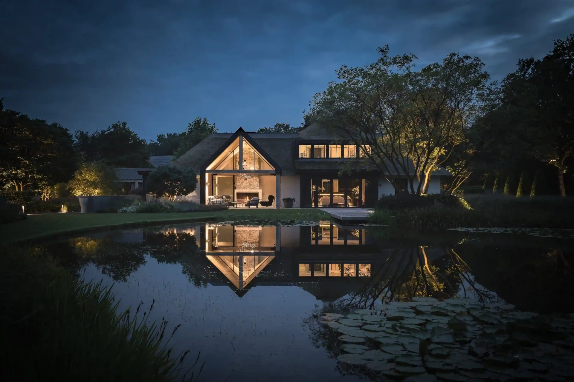Een panoramisch avondbeeld van een luxe rietgedekte villa door ERIC KANT, met een verlichte glazen vide, omringd door een parkachtige tuin en een grote vijver met lelies die het huis perfect weerspiegelt.