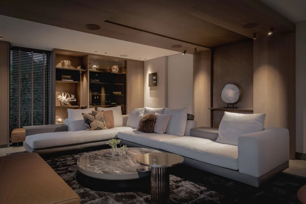 In de avonduren komt het verfijnde lichtplan van Villa Essenza volledig tot zijn recht. ERIC KANT creëert een warme, bijna filmische sfeer door gebruik te maken van subtiele spots en geïntegreerde LED-verlichting in de houten plafonds en maatwerk kasten. De royale hoekbank in een lichte tint contrasteert prachtig met het donkere, diepe tapijt en de marmeren salontafels. Decoratieve objecten, zoals het verlichte koraal en de artistieke sculptuur aan de wand, worden door de gerichte verlichting als kunstwerken gepresenteerd. Dit high-end interieur ademt rust en exclusiviteit, waarbij de natuurlijke materialen zoals hout en steen door het warme licht nog meer tot leven komen. Design door ERIC KANT.