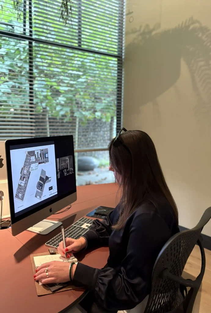Een lid van het team van ERIC KANT werkt geconcentreerd aan een bureau in de studio in Breda, met een gedetailleerde architectonische plattegrond op een groot iMac-scherm en handgeschreven aantekeningen in een schetsboek.