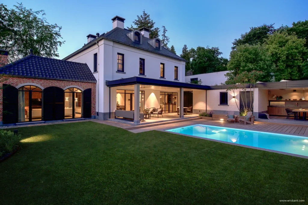 Een sfeervol verlichte Manor Villa bij avond door ERIC KANT, met een modern overdekt terras, een verlicht zwembad, een klassiek bijgebouw van baksteen en een strak gazon onder een diepblauwe lucht.