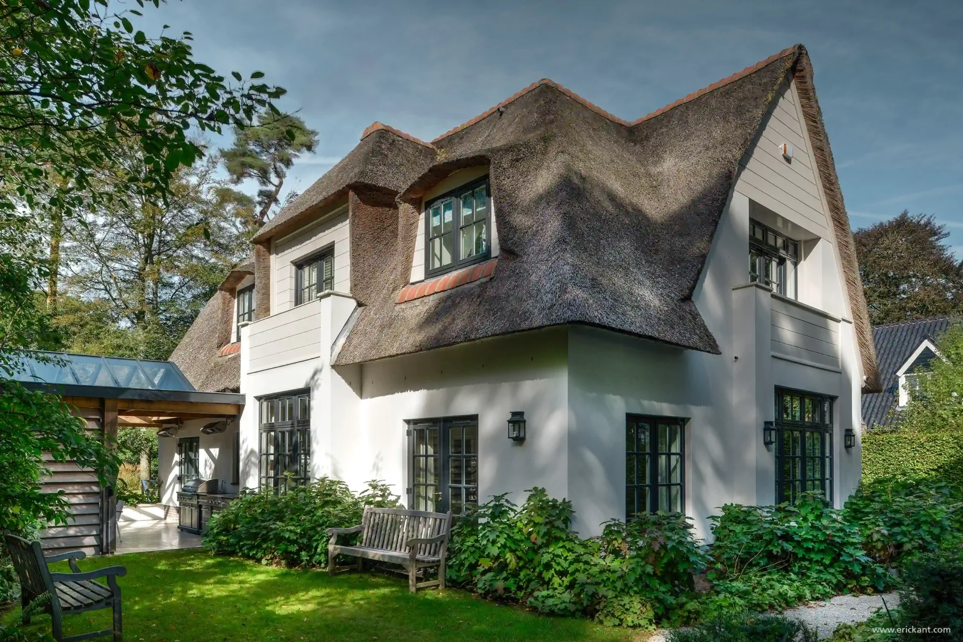 Een zijaanzicht van een klassieke witgepleisterde villa met een rieten kap in Laren door ERIC KANT, voorzien van een moderne glazen aanbouw met buitenkeuken en een verzorgde tuin.