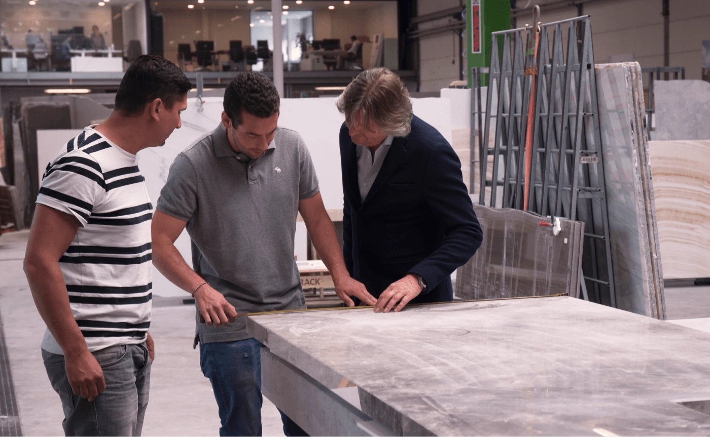 Eric Kant in overleg met vakmensen in een natuursteenwerkplaats, terwijl ze nauwkeurig een groot marmeren of stenen blad opmeten voor een maatwerkproject.
