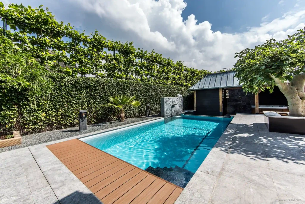 Een totaaloverzicht van een luxe wellness-tuin door ERIC KANT met een zwembad voorzien van waterval, een houten poolhouse, een jacuzzi en een rietgedekte villa op de achtergrond.