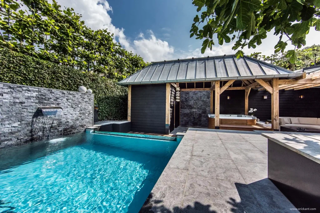 Een luxe wellness-tuin ontworpen door ERIC KANT met een azuurblauw zwembad inclusief waterval, een natuurstenen wand, een houten poolhouse met jacuzzi en een modern terras.
