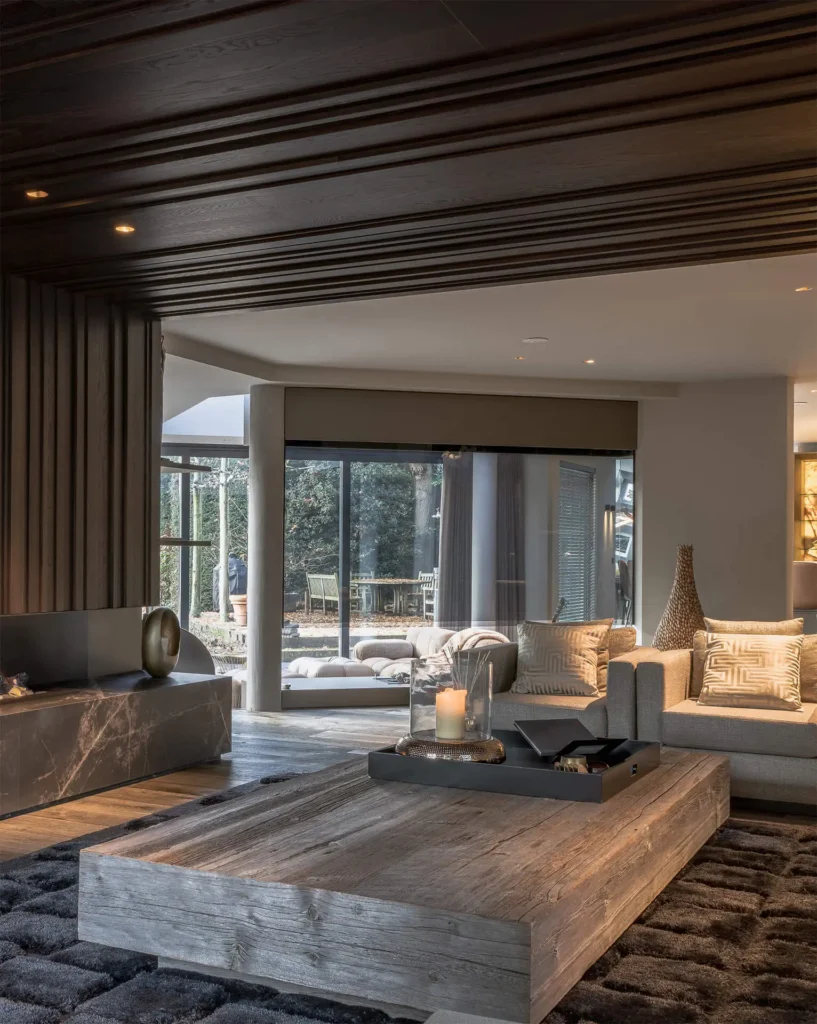 Een sfeervol high-end interieur ontworpen door ERIC KANT, met een robuuste houten salontafel, een luxe zithoek, een marmeren haardplateau en een karakteristiek houten lattenplafond dat doorloopt naar buiten.
