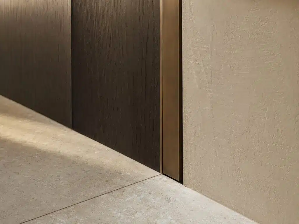 Aansluitdetails waar donker hout, een bronzen profiel, stucwerk en een natuurstenen vloer elkaar raken