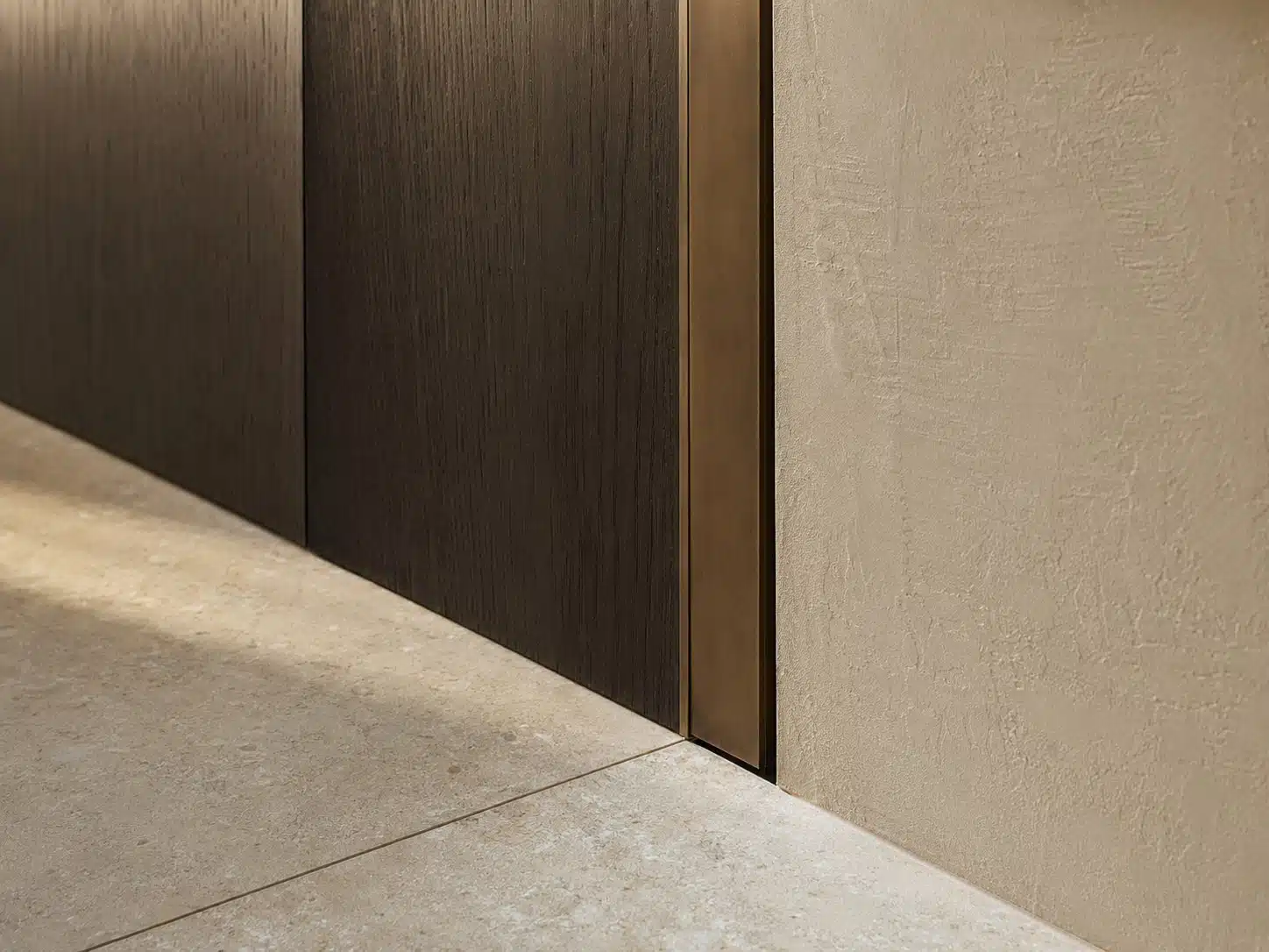 Aansluitdetails waar donker hout, een bronzen profiel, stucwerk en een natuurstenen vloer elkaar raken